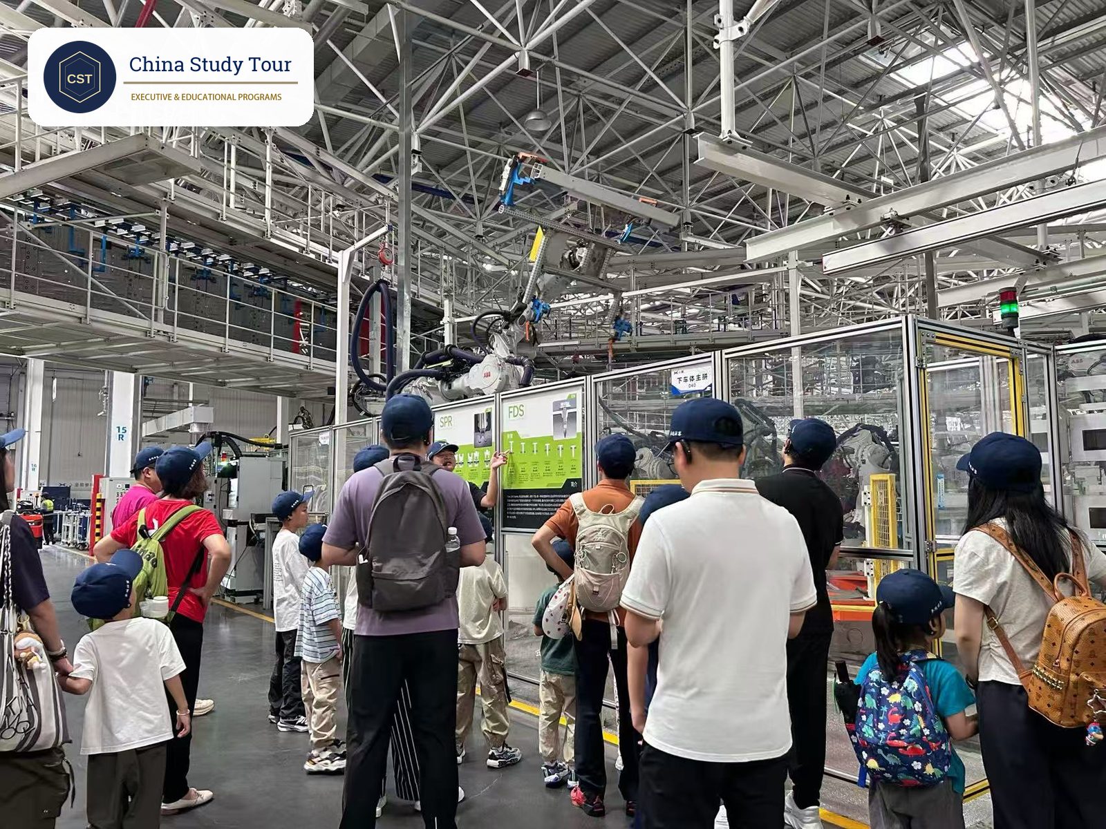 Students in matching caps and accompanying adults listen to a host guide beside an EV body-welding line